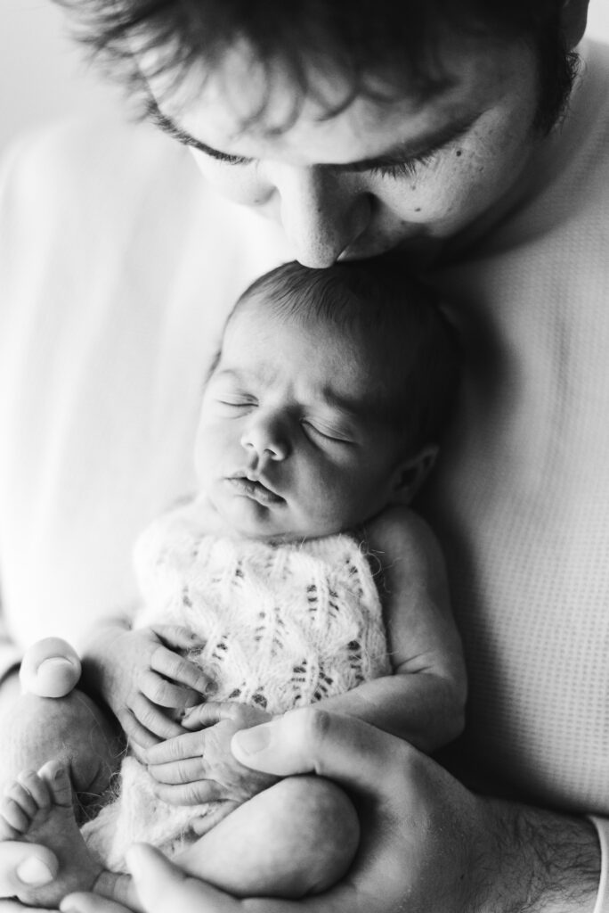 Dad holding his newborn baby girl and kissing her on the head at newborn photoshoot in West Sussex