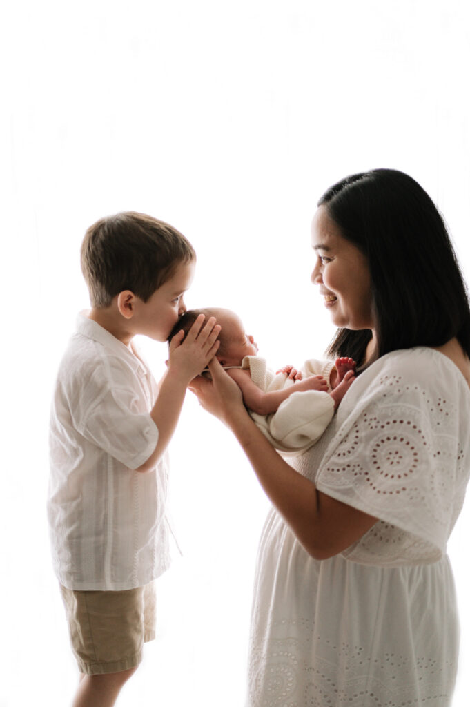 A mum holding her daughter and her little boy kissing his baby sisters head at Billingshurst newborn photoshoot