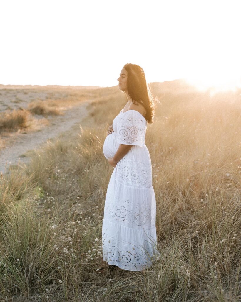 Pregnant woman in a white dress at the beach standing on long green grass with the sunset behind her at maternity photoshoot in Horsham