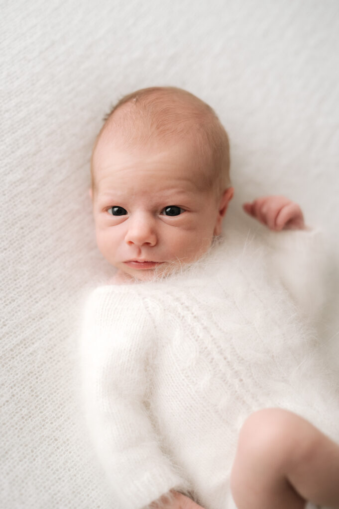 Newborn baby boy laying on a white soft blanket staring into the camera during a photoshoot in Horsham