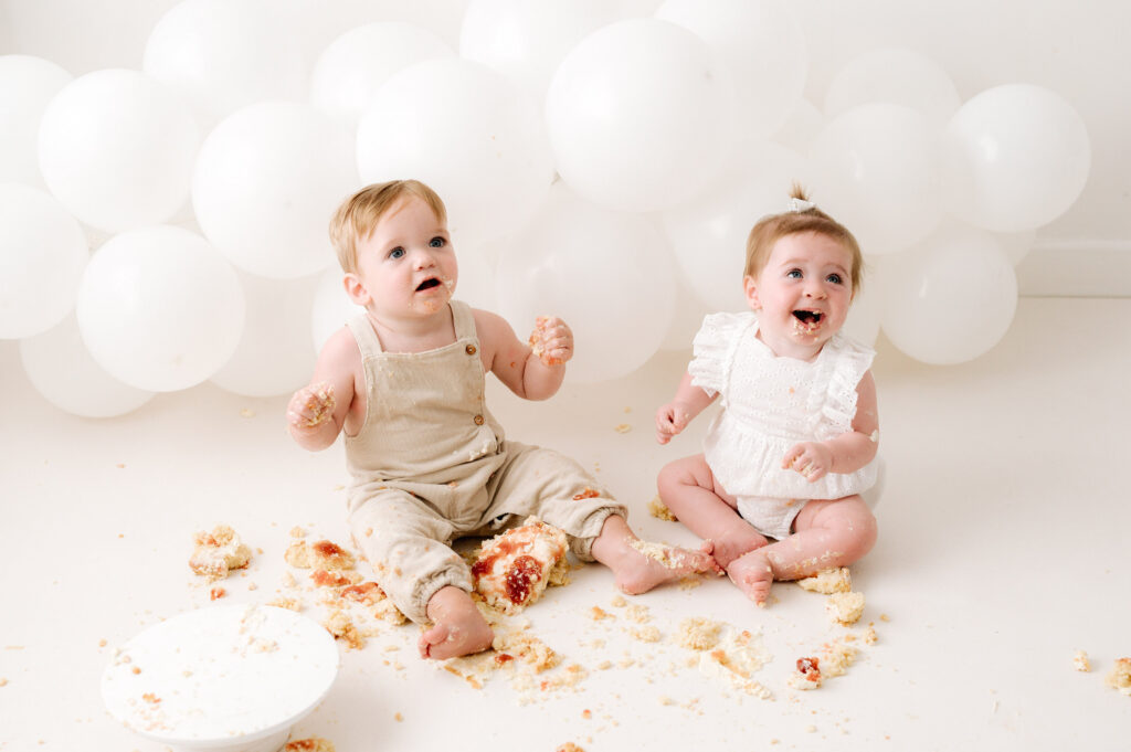 Twin 1 year olds smiling whilst eating a cake at Horsham cake smash photoshoot